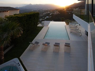 Großer Infinity-Pool mit heller Holzterrasse, mehreren Sonnenliegen und separatem Whirlpool, umrahmt von einer Gebirgslandschaft bei Sonnenuntergang.