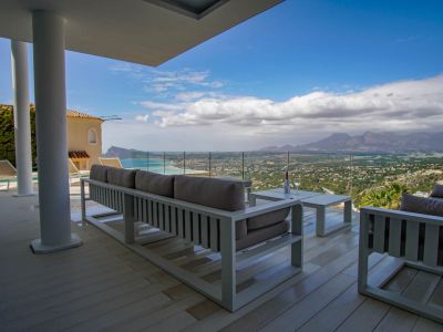 Großzügige überdachte Terrasse mit hellem Bodenbelag und weißen Loungemöbeln, die durch eine Glasbrüstung einen Panoramablick auf Meer, Küste und Berge bietet.