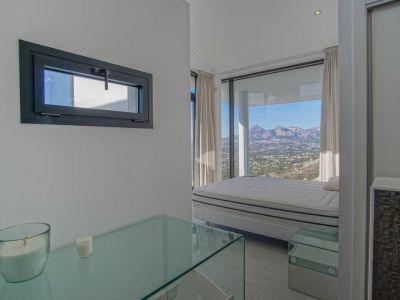 Helles Schlafzimmer mit Bett vor raumhohen Fenstern, die einen Panoramablick auf Berge und Tal bieten. Eine Glaskonsole und ein horizontales Fenster mit dunklem Rahmen sind ebenfalls sichtbar.