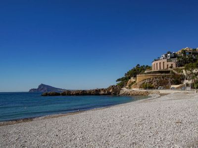 Kieselstrand mit klarem türkisfarbenem Wasser und felsiger Küste. Moderne Gebäude mit reflektierenden Fassaden und ein Rundbau säumen den Hang unter einem strahlend blauen Himmel.