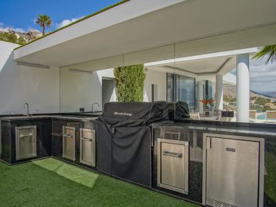 L-förmige Außenküche mit schwarzen Granitarbeitsplatten, Doppelspülbecken, überdachtem Grill und integrierten Edelstahlgeräten, mit reflektiertem Poolblick.