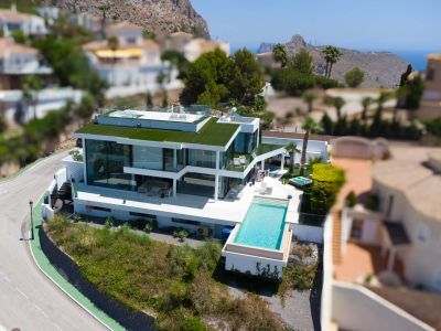 Luftaufnahme einer modernen weißen Villa mit Infinity-Pool, großen Glasfronten, begrüntem Dach und separatem Whirlpool, mit Berg- und Meerblick.