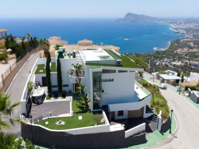 Luftaufnahme einer modernen weißen Villa mit mehrstufiger Architektur, begrünten Flachdächern und externem Glasaufzug. Das Anwesen umfasst eine private Auffahrt und bietet Panorama-Meer- und Küstenblicke.