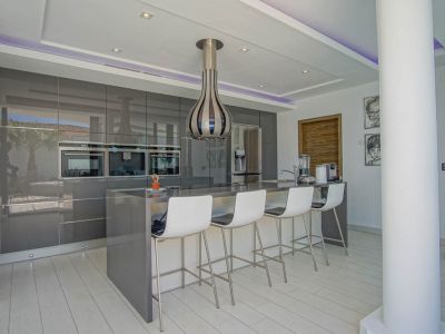 Moderne Küche mit hochglänzenden grauen Schränken, integrierten Öfen und einer Kücheninsel mit Sitzgelegenheiten für vier Personen. Indirekte LED-Beleuchtung im Deckenbereich.