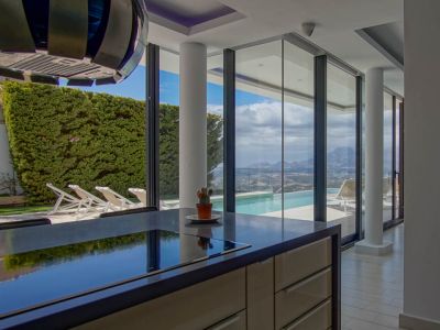 Moderne Kücheninsel mit dunkler Arbeitsplatte und integriertem Kochfeld. Die bodentiefe Fensterfront bietet einen weiten Blick auf den Infinity-Pool, das Tal und die Berge.