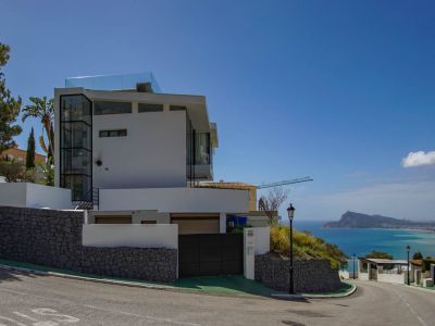 Moderne weiße Villa mit sichtbarem Glasaufzug und Dachterrassen-Brüstung. Steinmauern und ein automatisches Tor ergänzen den Panoramablick auf das Meer und die Küste.