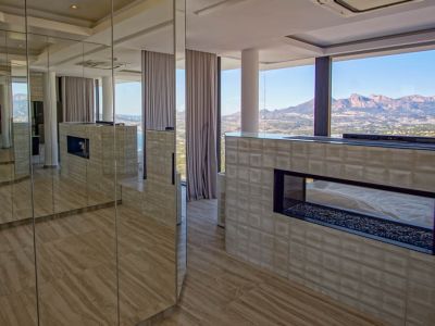 Moderner Innenraum mit transparentem Gaskamin und Kiesbett, in eine strukturierte Wand integriert. Spiegelpaneele reflektieren den Raum und die weiten Berg- und Meerblicke durch raumhohe Fenster.
