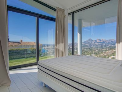 Modernes Schlafzimmer mit großem Bett und weiten Fenstern. Es bietet Panoramablicke auf das ferne Meer, eine grüne Rasenfläche und schroffe Berge unter klarem Himmel.