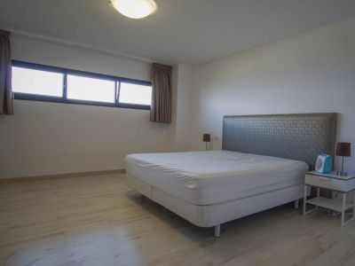 Schlafzimmer mit hellem Holzfußboden und weißen Wänden. Es verfügt über ein Bett mit strukturiertem Kopfteil, Nachttisch und ein horizontales Fenster mit dunklen Rahmen.