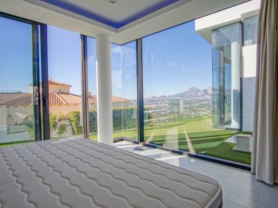 Schlafzimmer mit raumhohen Eckglaswänden, das einen Panoramablick auf ferne Berge, ein Tal und benachbarte Dächer bietet. Ein weißer Stützpfeiler und direkter Zugang zu Kunstrasen sind vorhanden.