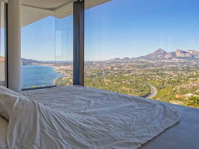 Schlafzimmer mit raumhohen Glasfenstern und Panoramablick auf das Mittelmeer, eine Küstenstadt und ferne Berge. Ein niedriges Bett mit weißer Bettwäsche orientiert sich zur Aussicht.