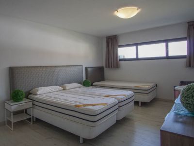 Schlafzimmer mit zwei kombinierten Einzelbetten, grauen strukturierten Kopfteilen, einem dritten Einzelbett im Hintergrund, hellem Holzboden und horizontalen Fenstern.