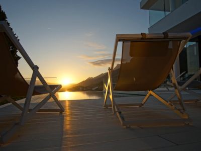 Zwei moderne Liegestühle auf heller Terrasse blicken auf einen Infinity-Pool, der den goldenen Sonnenuntergang über den Bergen spiegelt.