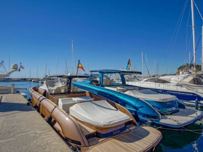 Zwei moderne Motorboote, eines bronze mit Teakdeck und ein weiteres metallicblau, liegen in einem Yachthafen unter klarem Himmel. Spanische Flaggen sind unter den Yachten im Hintergrund zu sehen.