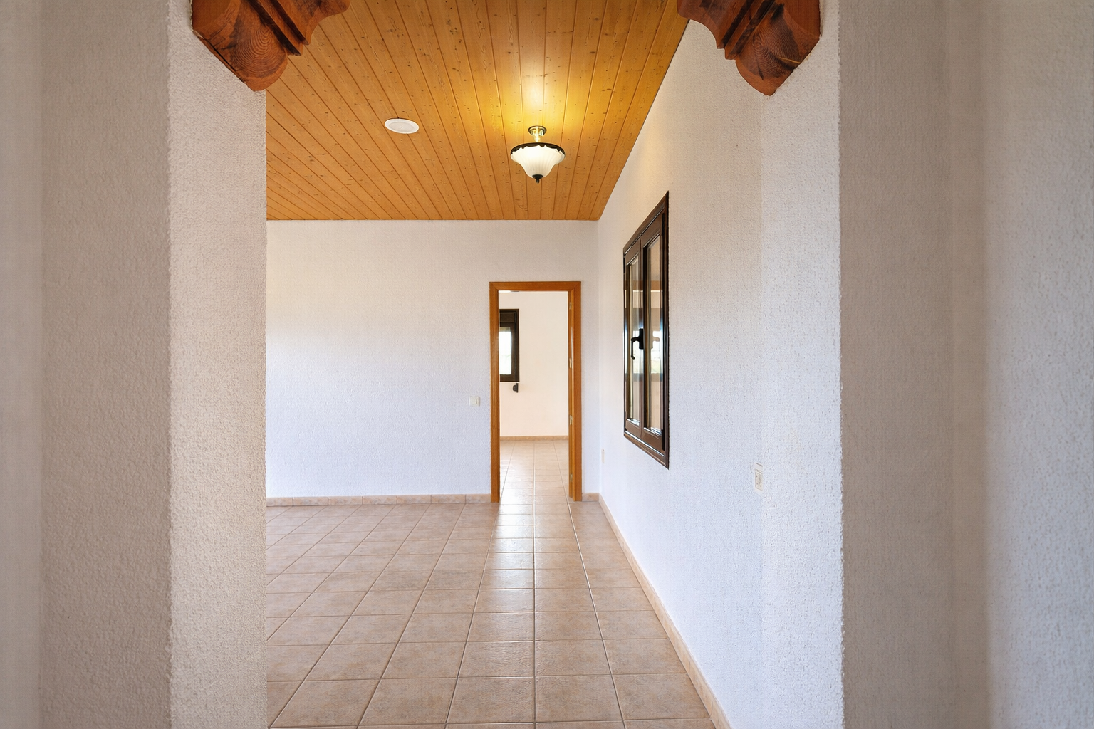 Innenflur mit hellen Fliesenböden und strukturierten weißen Wänden. Holzdeckenpaneele mit Deckenleuchte und geschnitzten Holzelementen am Bogen.