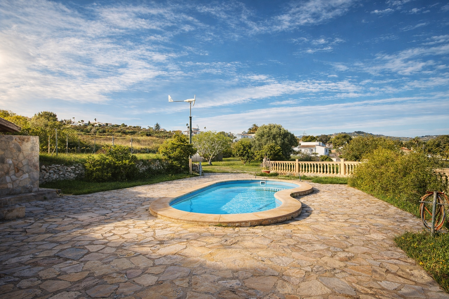 Organisch geformter Pool auf einer weitläufigen Terrasse aus unregelmäßigen Steinplatten. Grüne Hügel, Vegetation und eine kleine Windturbine im Hintergrund sichtbar.