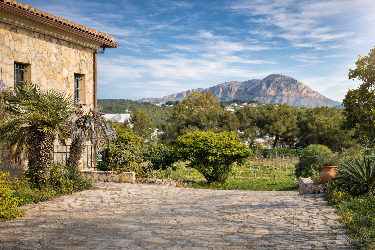 Rustikale Steinfassade der Finca mit Palmen, gepflasterter Zufahrt und Blick auf den Montgó, umgeben von Weinbergen und mediterraner Vegetation.