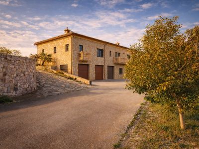 Die steinerne Außenfassade der Finca zeigt drei integrierte Garagen und elegante Balkone, zugänglich über eine gepflasterte Zufahrt und eine geschwungene Steinmauer.