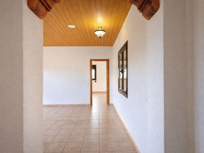 Innenflur mit hellen Fliesenböden und strukturierten weißen Wänden. Holzdeckenpaneele mit Deckenleuchte und geschnitzten Holzelementen am Bogen.