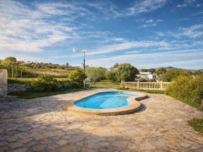 Organisch geformter Pool auf einer weitläufigen Terrasse aus unregelmäßigen Steinplatten. Grüne Hügel, Vegetation und eine kleine Windturbine im Hintergrund sichtbar.