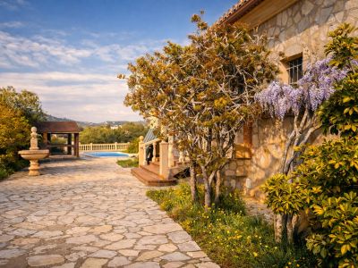 Steinfassade der Finca mit blühendem Blauregen. Ein gepflasterter Weg führt zum Poolbereich und einer Holzpergola, mit einem Steinbrunnen im Garten.