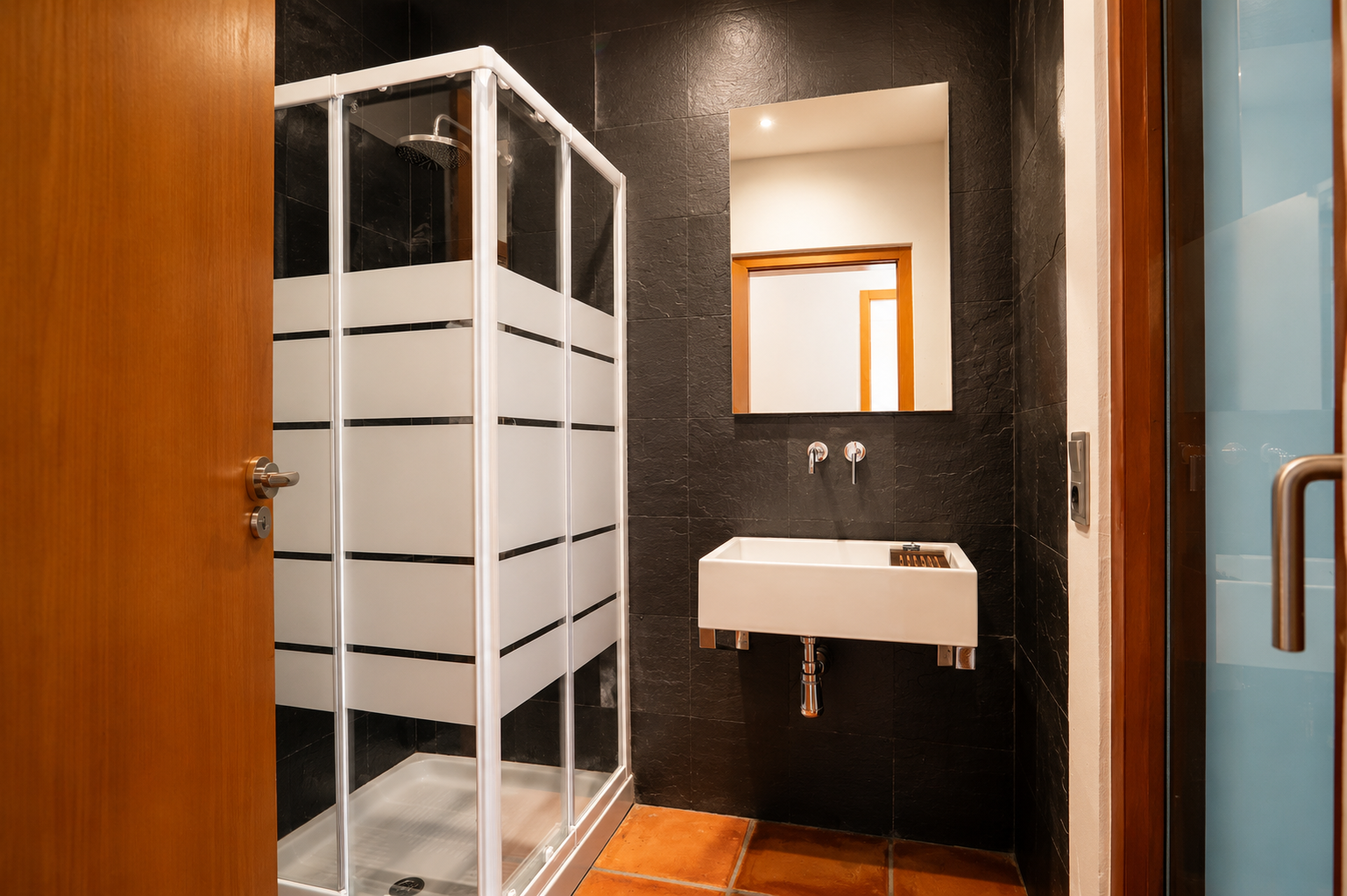 Badezimmer mit Duschkabine aus Milchglasstreifen, rechteckigem Wandwaschbecken und dunklen strukturierten Wandfliesen. Terrakotta-Bodenfliesen ergänzen den Raum.