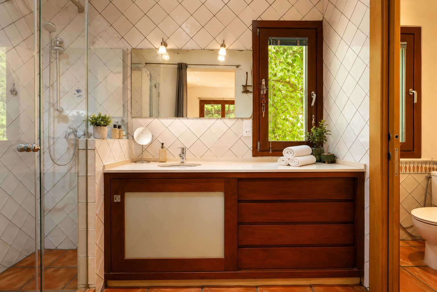 Badezimmer mit Glasdusche, dunklem Holzwaschtisch und weißer Arbeitsplatte. Ein Fenster bietet Blick ins Grüne, ergänzt durch rautenförmige weiße Fliesen und Terrakottaböden.
