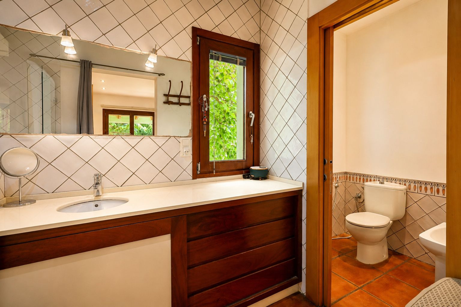Badezimmerwaschtisch mit hellem Becken und dunklem Holzunterschrank. Ein großer Spiegel mit Wandleuchten und ein Holzfenster bieten Blick auf üppiges Grün im Freien.