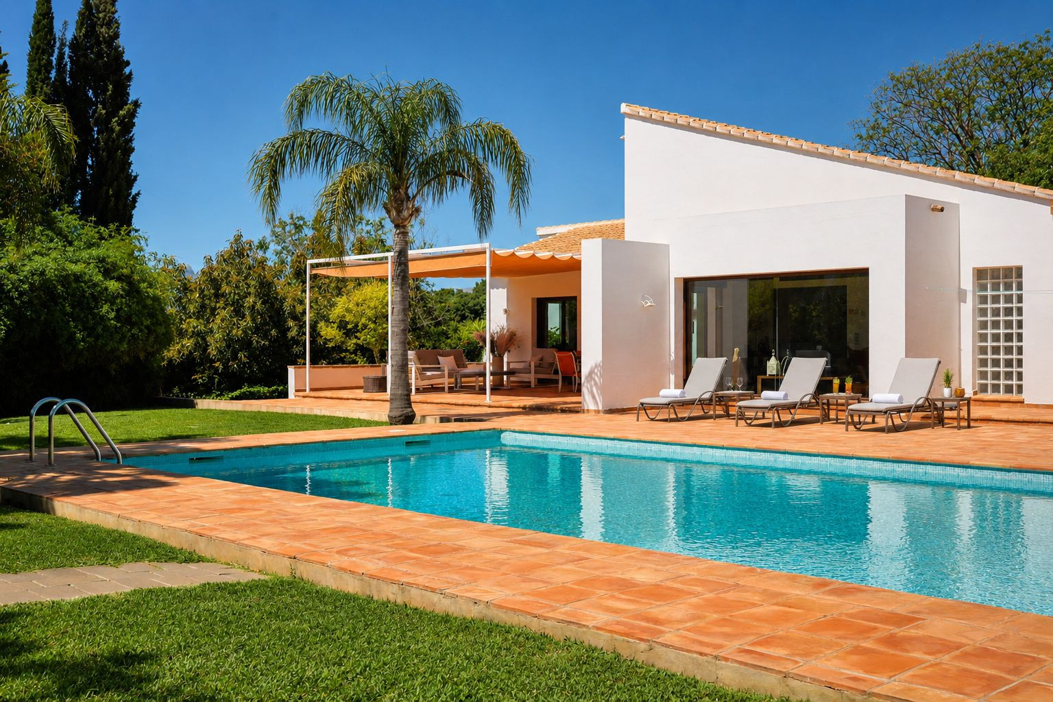 Blau gefliester Pool mit Terrakotta-Umfassung und Rasenfläche. Sonnenliegen stehen vor der weißen Hauptfassade, neben einer schattigen Pergola mit Lounge-Möbeln.