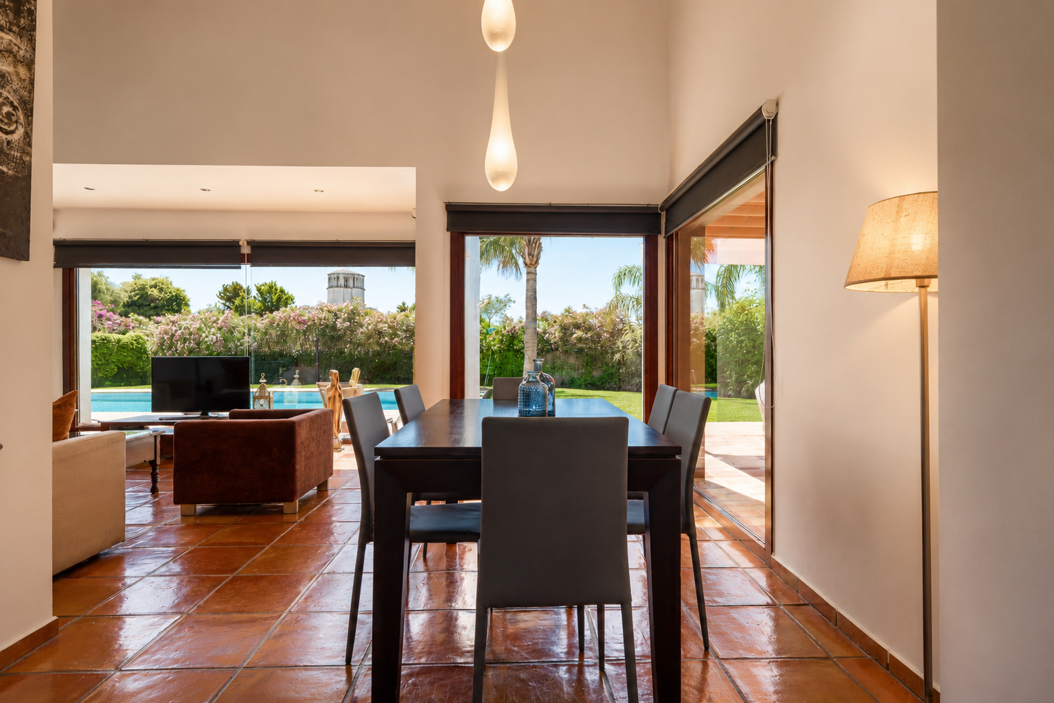 Der Essbereich mit dunklem Holztisch und Terrakottafliesen wird von Tageslicht durch große Glastüren erhellt. Er bietet direkten Blick auf den Garten, den Pool und einen entfernten Turm.