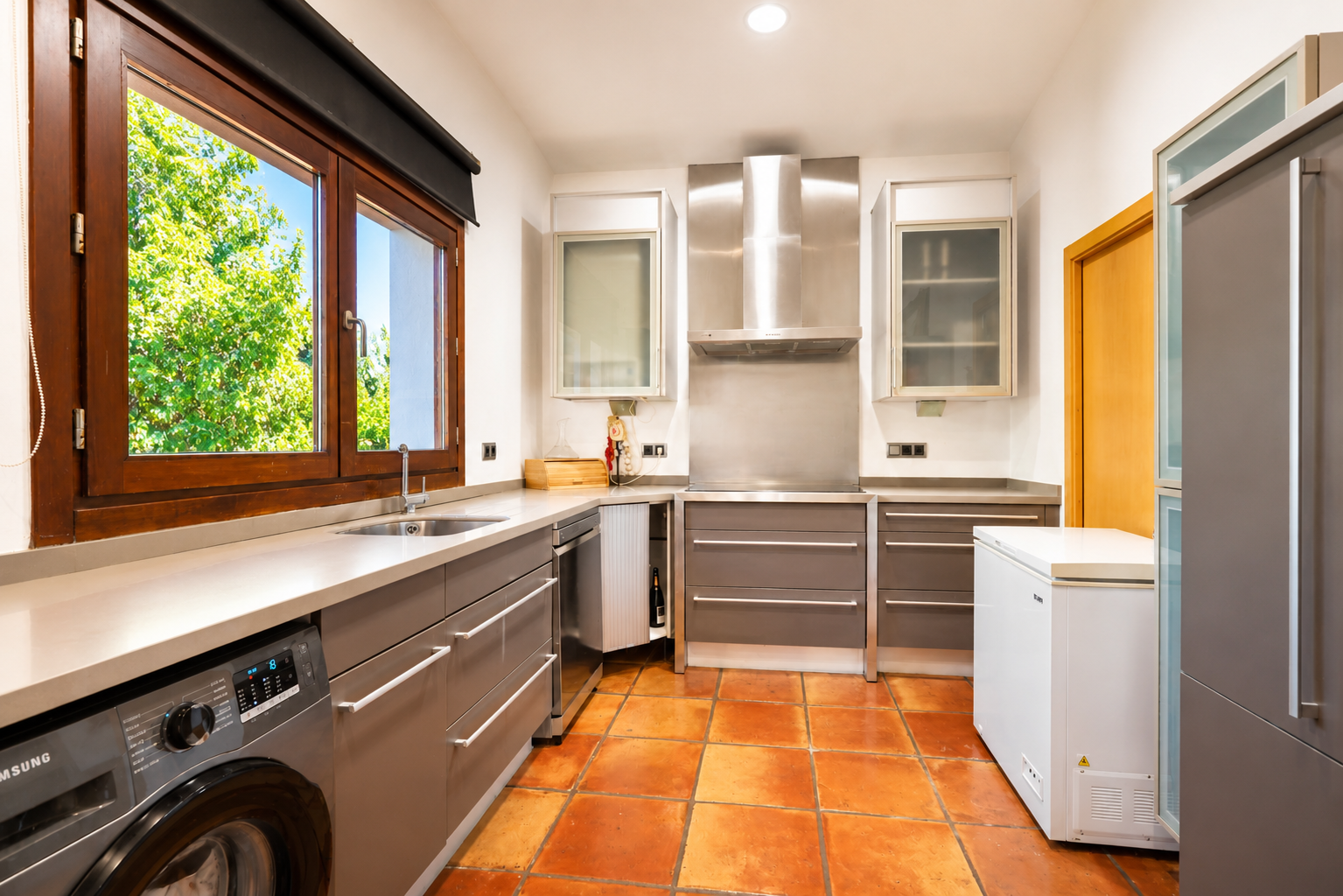 Funktionale Küche mit integrierter Waschmaschine, handgefertigtem Terrakotta-Boden und großem Holzfenster mit Gartenblick.