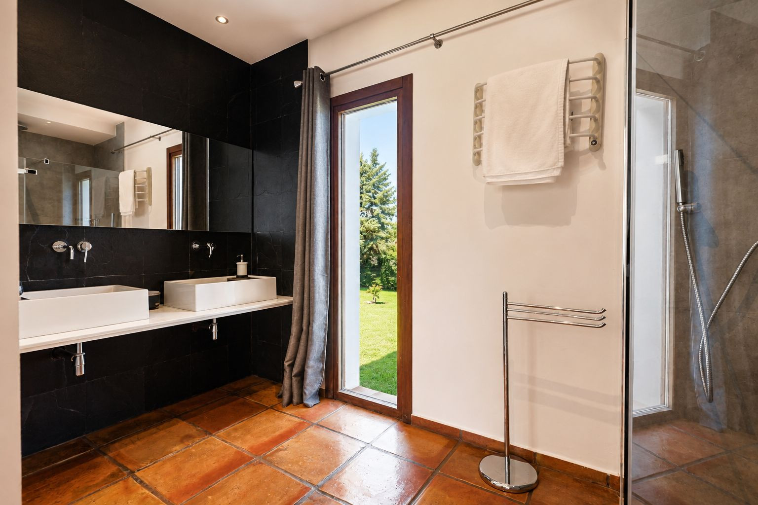 Geräumiges Badezimmer mit Terrakotta-Bodenfliesen, Doppelwaschtisch mit weißen rechteckigen Waschbecken und schwarzer Fliesenwand. Großes Fenster mit Gartenblick und Glasdusche.