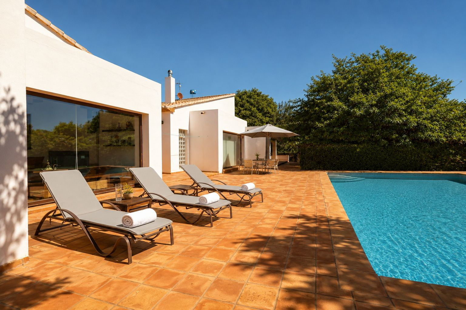 Großzügige Poolterrasse mit Terrakottafliesen, drei Sonnenliegen und einem schattigen Essbereich. Die weiße Fassade mit großen Fenstern spiegelt die grüne Umgebung wider.