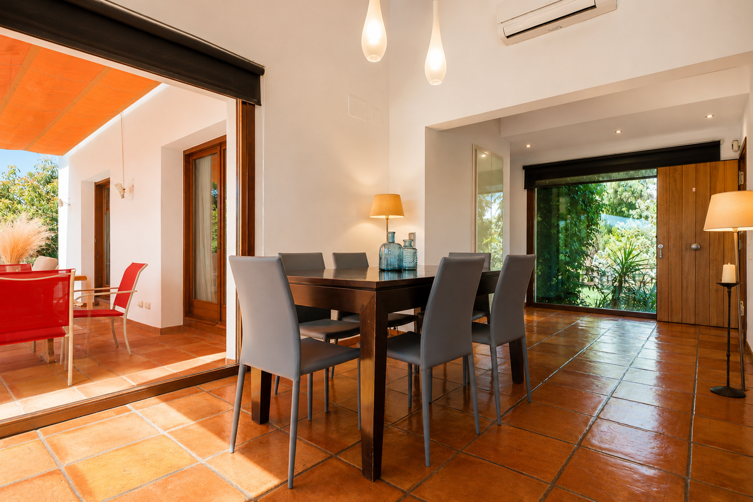 Heller Essbereich mit dunklem Holztisch und sechs grauen Stühlen auf Terrakottafliesen. Große Glastüren führen zu einer Außenterrasse mit orangefarbener Markise und Gartenblick.