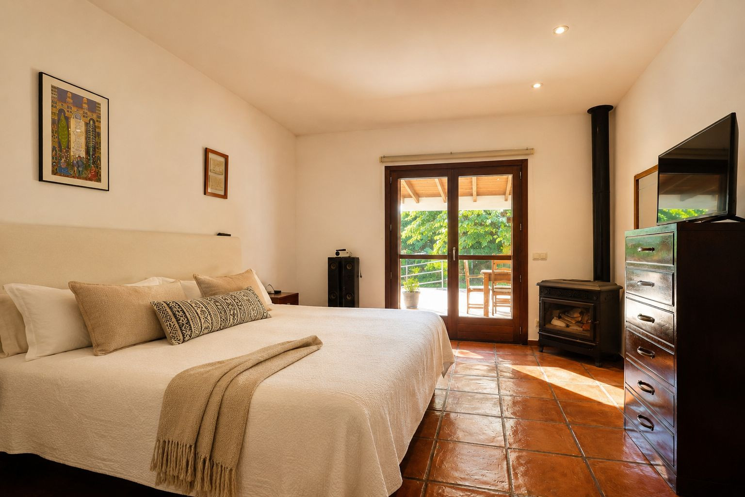Helles Schlafzimmer mit großem Bett, traditionellen Terrakotta-Fliesen und einem gemütlichen Holzofen. Doppelte Glastüren führen direkt auf eine von Grün umgebene Außenterrasse.