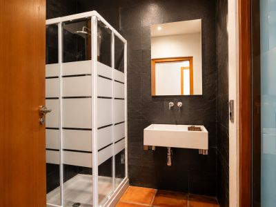 Badezimmer mit Duschkabine aus Milchglasstreifen, rechteckigem Wandwaschbecken und dunklen strukturierten Wandfliesen. Terrakotta-Bodenfliesen ergänzen den Raum.