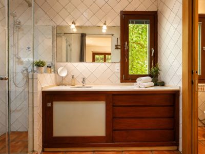 Badezimmer mit Glasdusche, dunklem Holzwaschtisch und weißer Arbeitsplatte. Ein Fenster bietet Blick ins Grüne, ergänzt durch rautenförmige weiße Fliesen und Terrakottaböden.