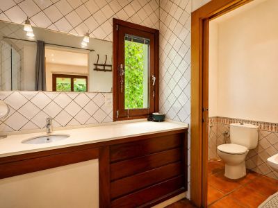 Badezimmerwaschtisch mit hellem Becken und dunklem Holzunterschrank. Ein großer Spiegel mit Wandleuchten und ein Holzfenster bieten Blick auf üppiges Grün im Freien.