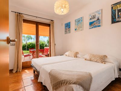 Doppelzimmer mit Terrakotta-Boden und direktem Zugang zur Außenterrasse. Die Glastür öffnet sich zu einem grünen Garten mit Palmen und roten Gartenmöbeln.