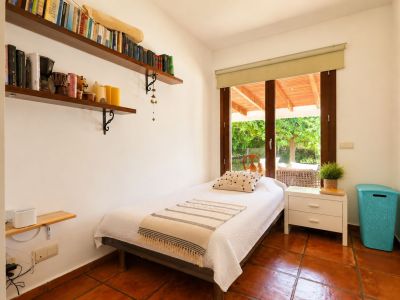 Einzelzimmer mit Terrakotta-Fliesenboden, weißen Wänden und direktem Zugang zu einer überdachten Terrasse durch eine große Holzglastür. Wandregale mit Büchern sind vorhanden.