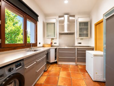 Funktionale Küche mit integrierter Waschmaschine, handgefertigtem Terrakotta-Boden und großem Holzfenster mit Gartenblick.