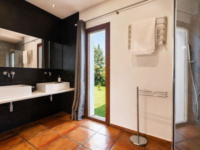 Geräumiges Badezimmer mit Terrakotta-Bodenfliesen, Doppelwaschtisch mit weißen rechteckigen Waschbecken und schwarzer Fliesenwand. Großes Fenster mit Gartenblick und Glasdusche.