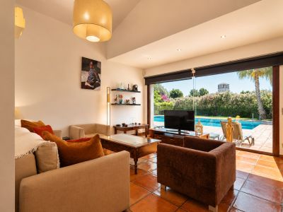 Geräumiges Wohnzimmer mit Terrakottafliesen und direktem Zugang zum Poolbereich. Helles Tageslicht durchflutet den Raum, der bequeme Sitzgelegenheiten und Gartenblick mit einem entfernten Turm bietet.