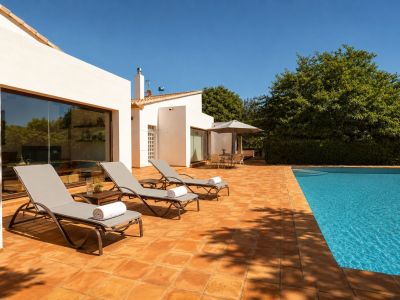 Großzügige Poolterrasse mit Terrakottafliesen, drei Sonnenliegen und einem schattigen Essbereich. Die weiße Fassade mit großen Fenstern spiegelt die grüne Umgebung wider.