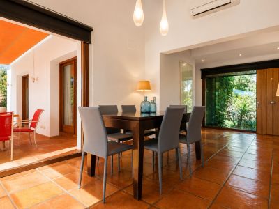 Heller Essbereich mit dunklem Holztisch und sechs grauen Stühlen auf Terrakottafliesen. Große Glastüren führen zu einer Außenterrasse mit orangefarbener Markise und Gartenblick.