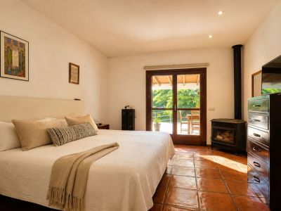 Helles Schlafzimmer mit großem Bett, traditionellen Terrakotta-Fliesen und einem gemütlichen Holzofen. Doppelte Glastüren führen direkt auf eine von Grün umgebene Außenterrasse.