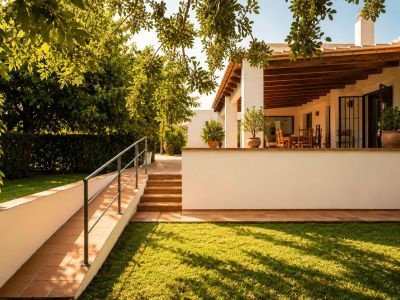 Überdachte Naya mit sichtbaren Holzbalken und Essbereich im Freien, mit Blick auf einen gepflegten Rasen und üppigen mediterranen Garten, zugänglich über Rampe und Treppen.