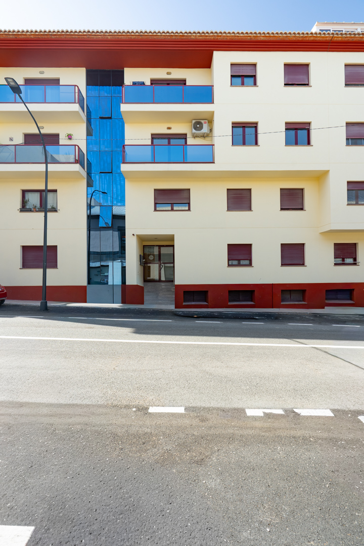 Außenansicht des Wohngebäudes mit cremefarbener Fassade, blauen Glasbalkonen und roten Rollläden. Ein zentraler blauer Glasabschnitt und ein ebenerdiger Eingang mit Rampe prägen das Erscheinungsbild.