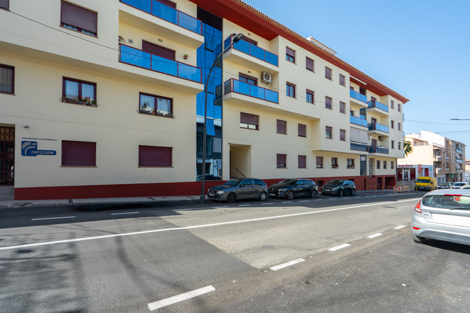 Außenansicht des Wohngebäudes mit heller Fassade, blauen Glasbalkonen, Fenstern mit weinroten Rollläden und einer vertikalen Glasfront.