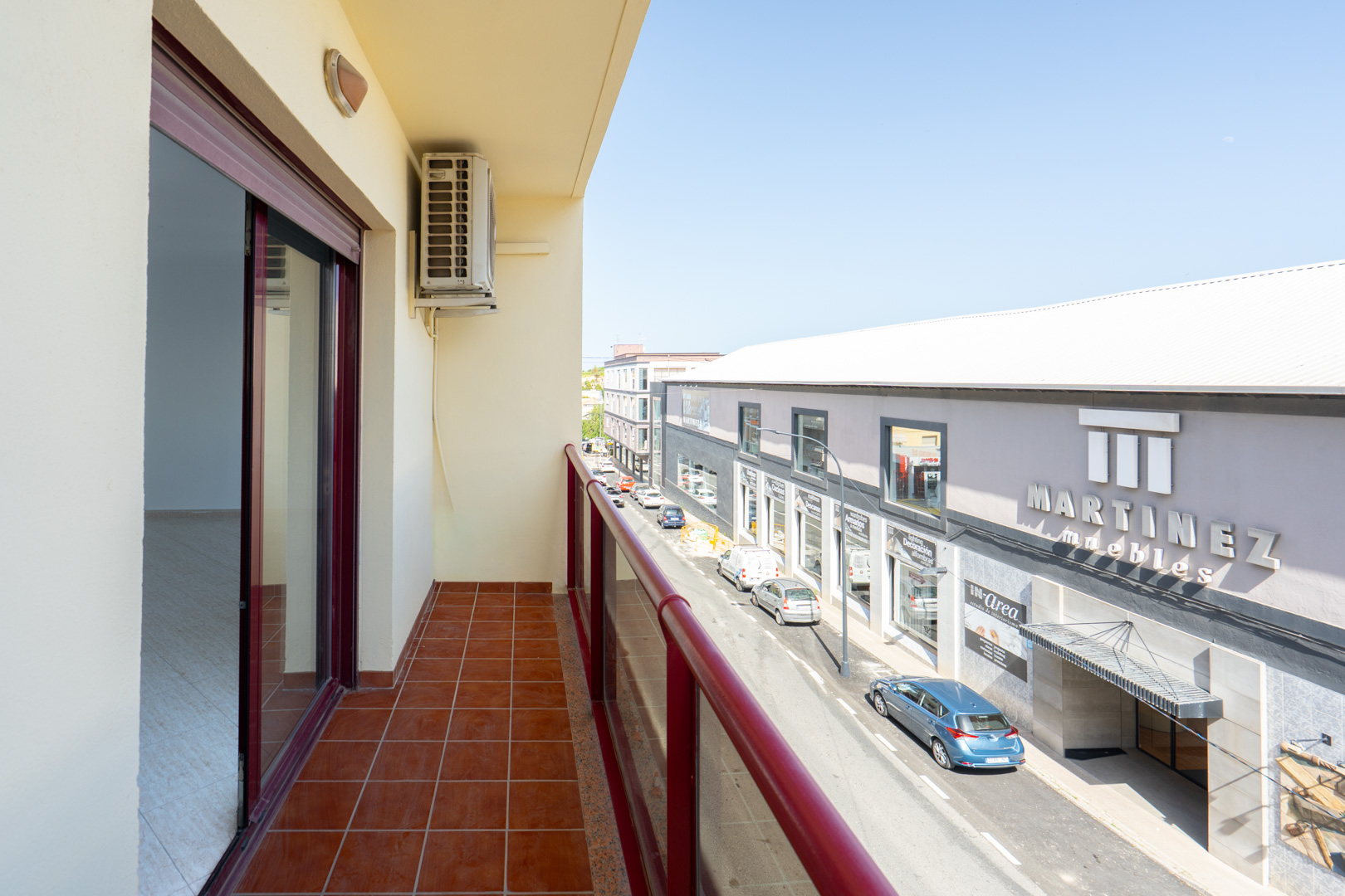 Außenbalkon mit Terrakottafliesen und rotem Geländer. Eine Klimaanlage ist sichtbar, mit Blick auf die Straße und das Geschäft "Martinez Muebles".